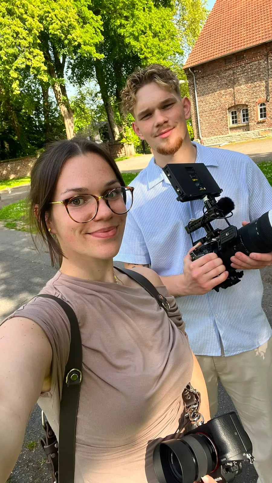 Selfie zweier Filmschaffender mit Kameras vor einem Backsteingebäude im Sonnenlicht.
