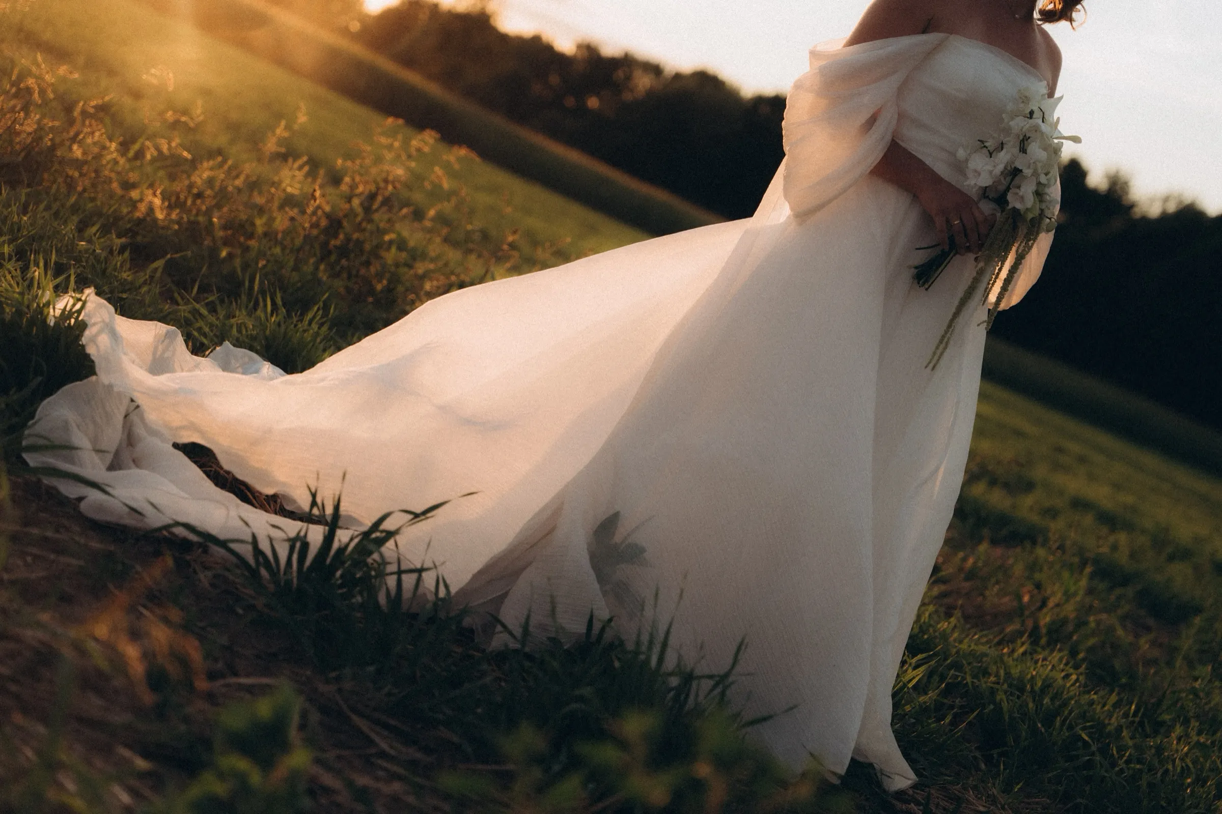 Die Schleppe des Brautkleids zieht durch hohes Gras, die Abendsonne steht flach dahinter.