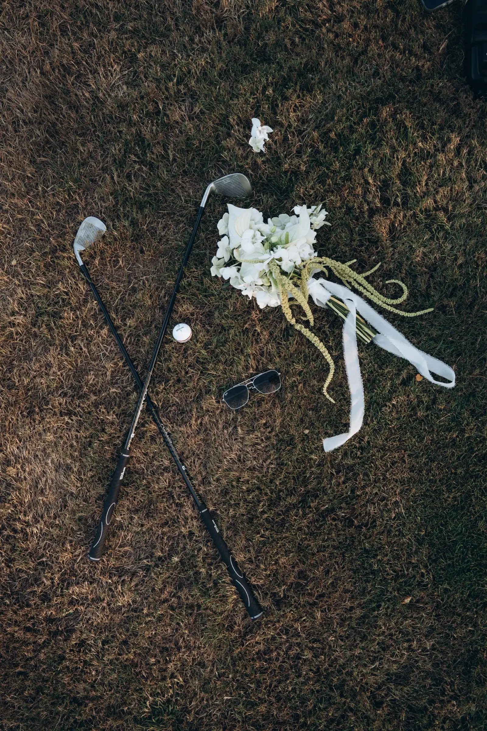 Zwei Golfschläger, ein Ball und der Brautstrauß liegen nebeneinander im Gras.